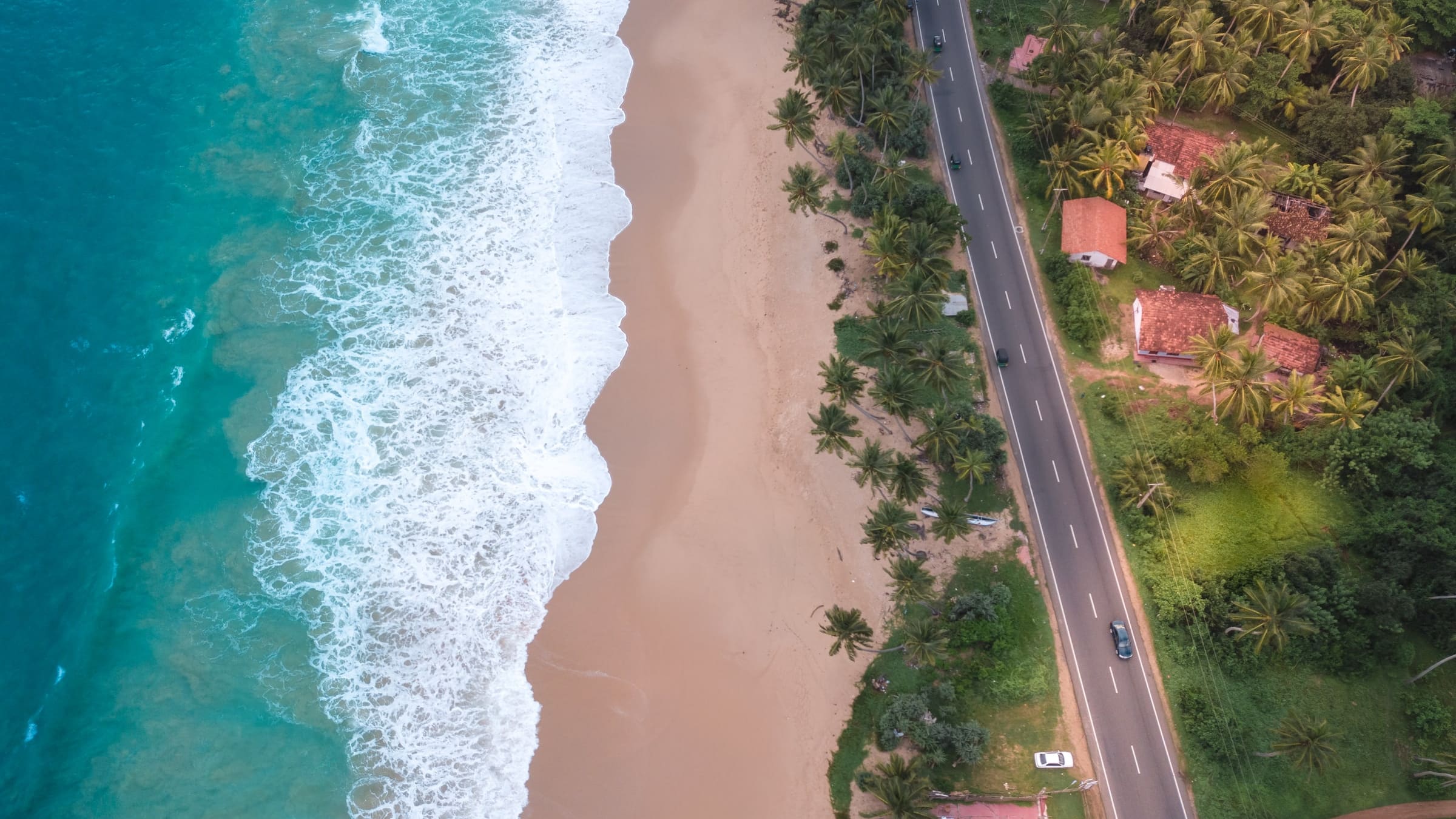 The beach at Aluthgama from the air, surf breaking along the sand beside the coast road and palms.