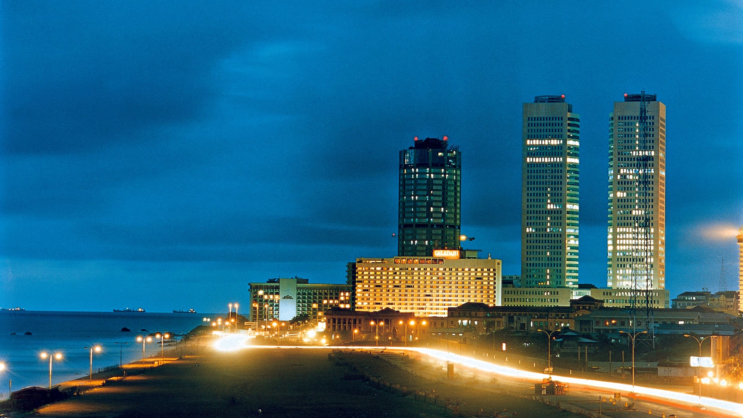 The Colombo sea front at dusk, lit towers along the shore reflected in the water.