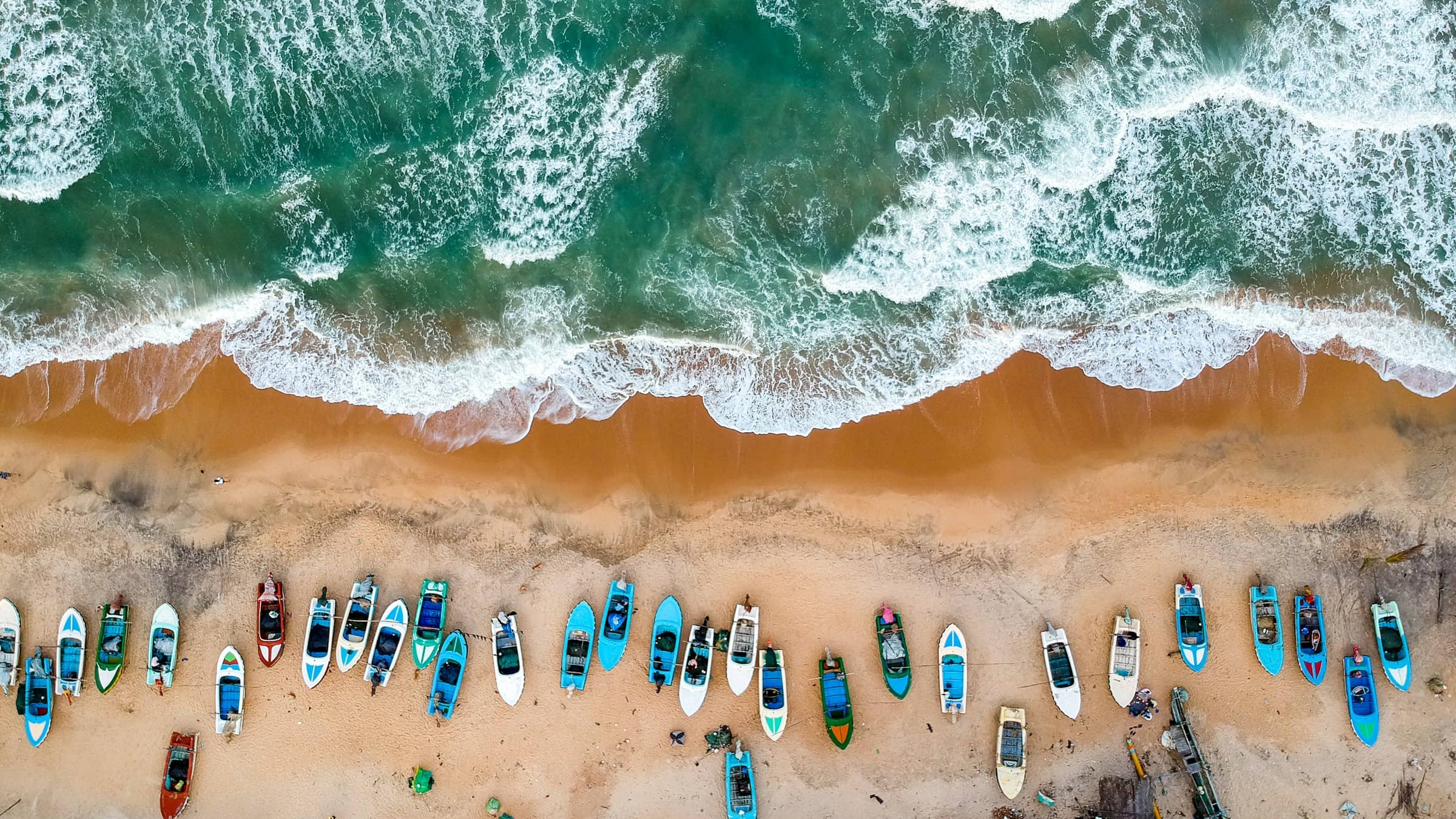 Painted fishing boats drawn up in a row on the sand, seen from directly above.
