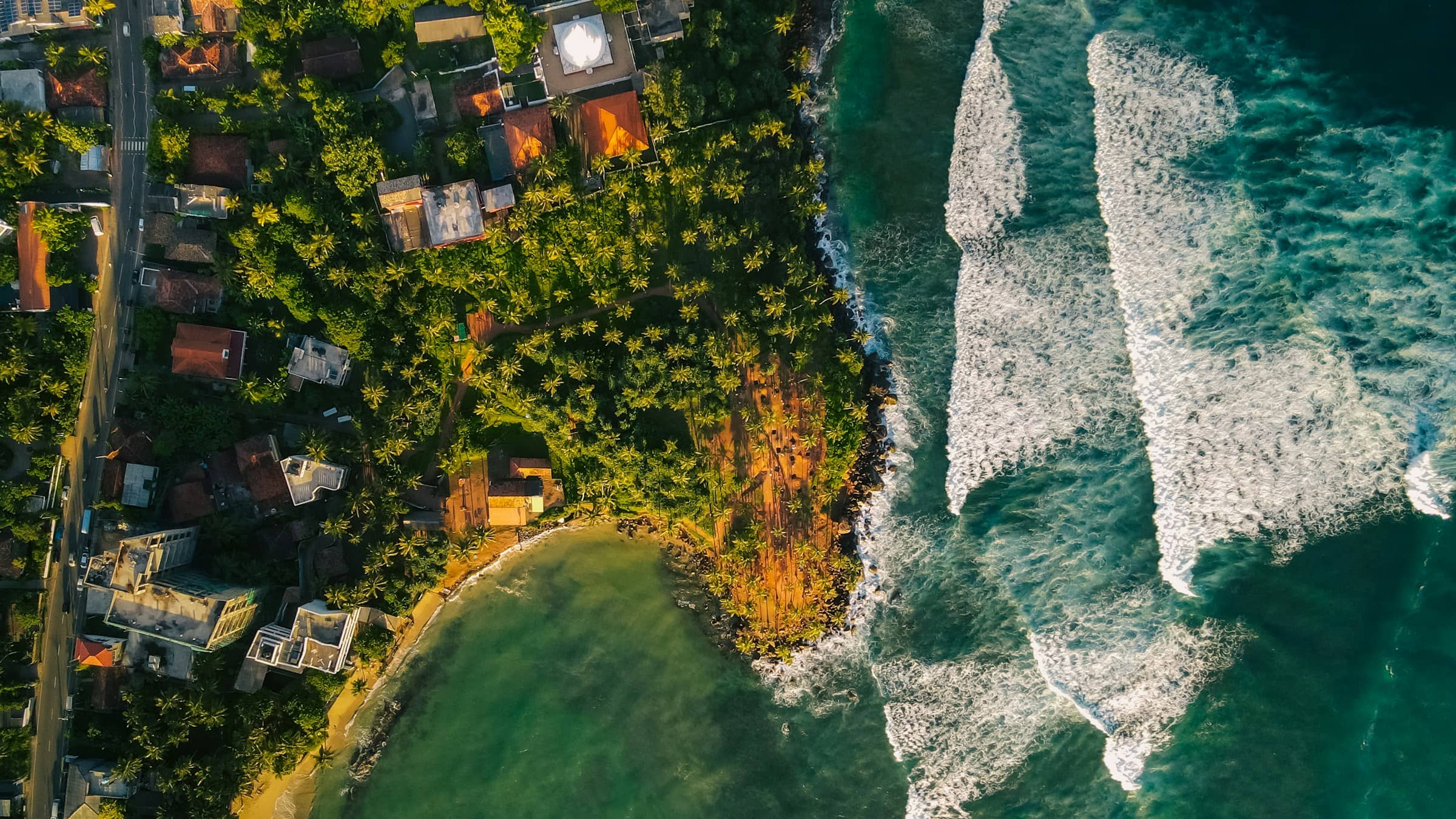A village on a green headland from the air, with surf breaking along both sides.