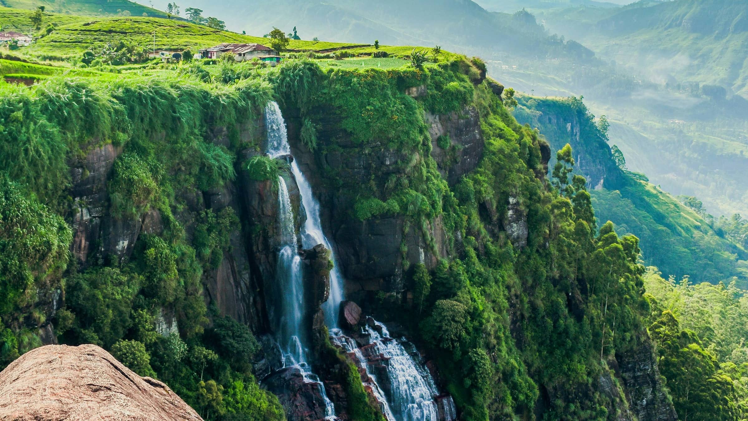 A waterfall dropping down a green cliff into a misty hill-country valley.