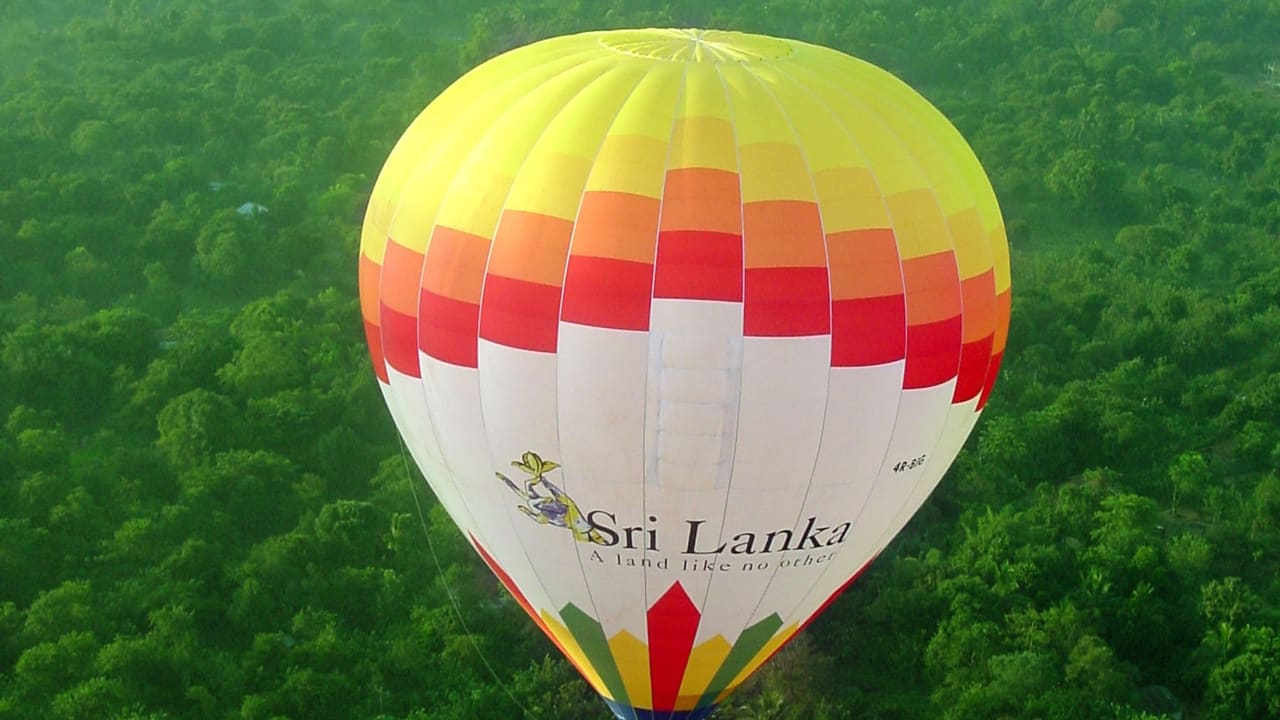 A hot air balloon drifting low over dense green dry-zone forest at first light.