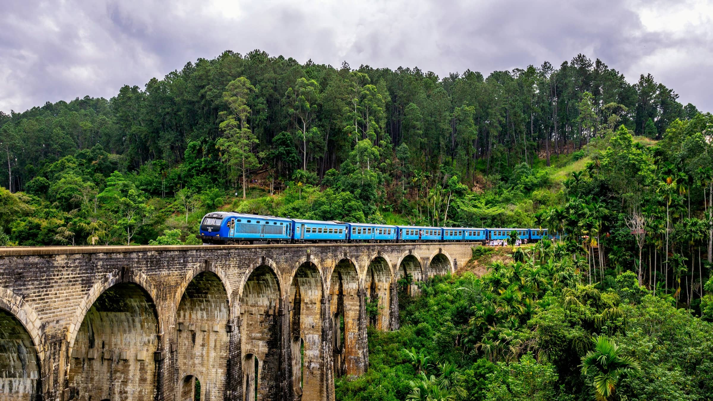 A blue train crossing the Nine Arch Bridge at Demodara, surrounded by dense hill-country jungle.