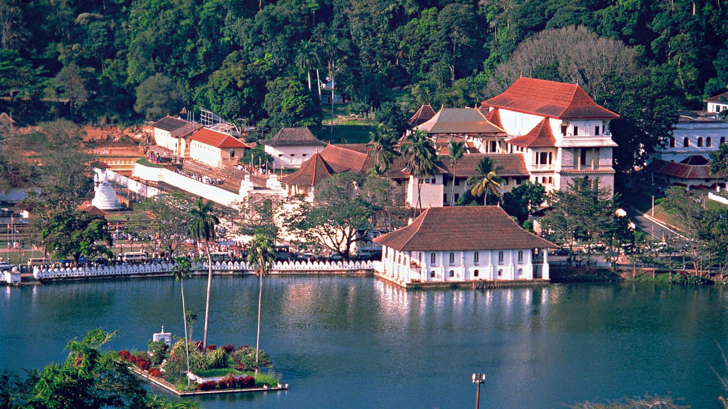 The Temple of the Sacred Tooth Relic across Kandy Lake, red-tiled roofs and white walls against forested hills.
